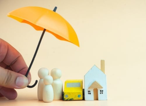 Hand holding a miniature yellow umbrella over wooden family, house and car - protection