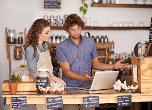 A young man and lady working behind a cafe counter looking at a laptop